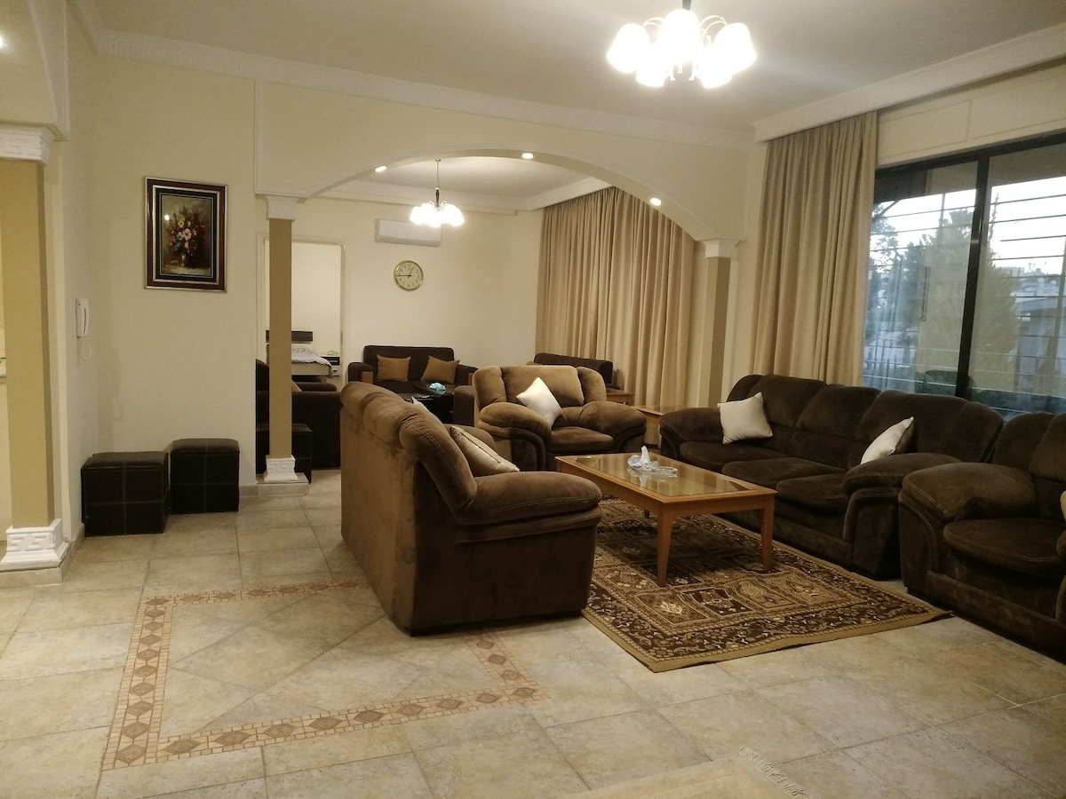 A spacious living area features comfortable brown sofas arranged around a wooden coffee table, placed on a decorative area rug. Large windows allow natural light to fill the room, complemented by soft curtains. Detailed wall decor and a clock add to the inviting ambiance.