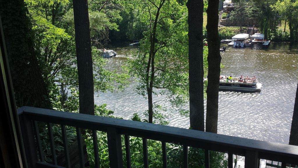 A view from a balcony showcases a tranquil waterway surrounded by lush greenery. Boats can be seen gliding along the water, framed by towering trees that provide shade and natural beauty.