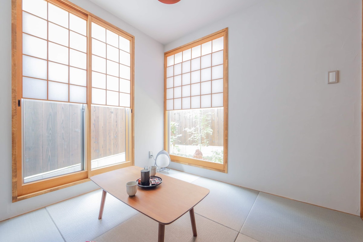 A light-filled room features two large windows with traditional latticework, allowing views of the garden. A low wooden table and simple seating on tatami mats provide a serene space. Soft natural light enhances the minimalistic decor, creating a calming ambiance.