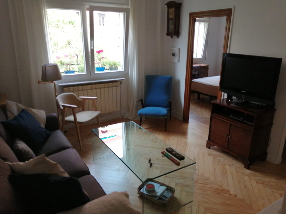 A comfortable sitting area features a brown sofa and two chairs positioned around a glass coffee table. A television is placed on a wooden cabinet, while a large window provides natural light and views of greenery. The room showcases wooden flooring and light-colored walls.