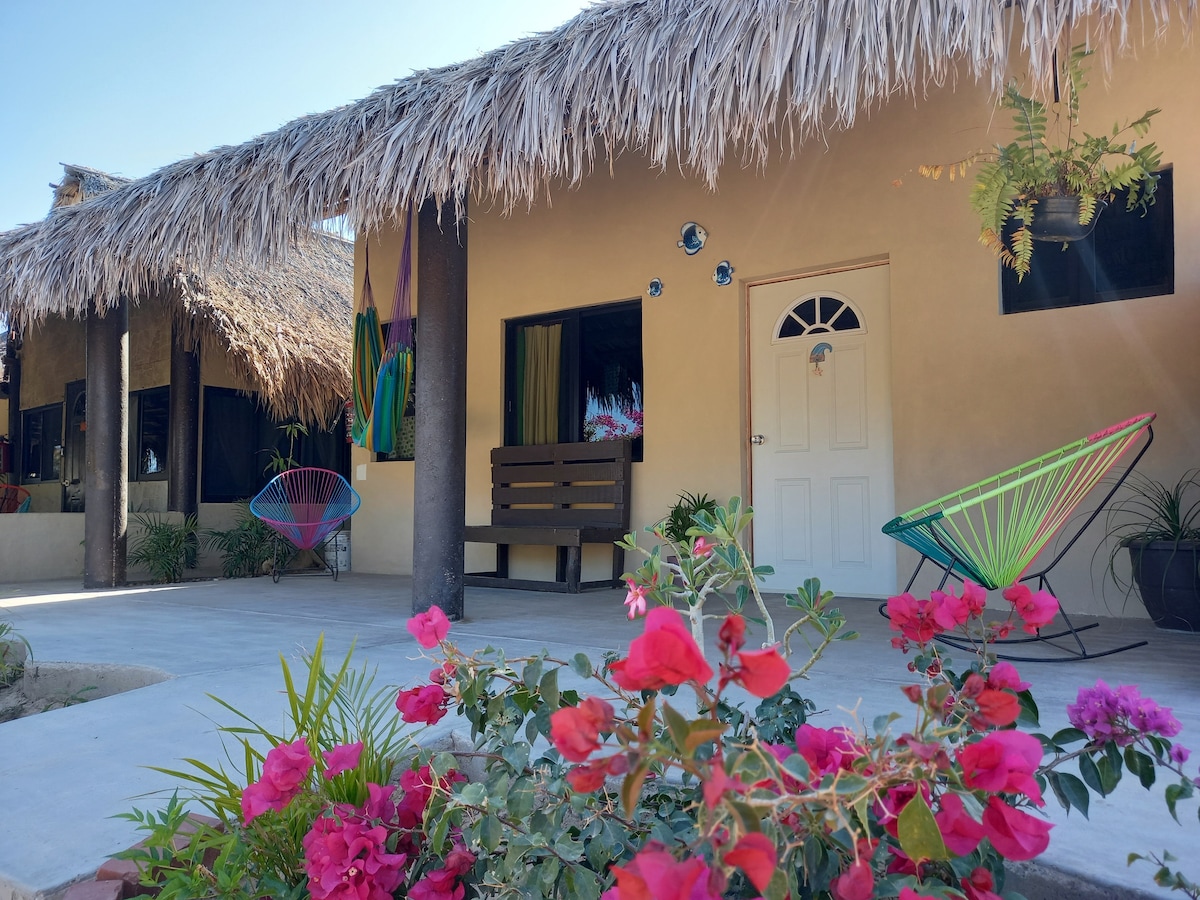A charming outdoor area features vibrant pink flowers in the foreground, while a pathway leads to a single-story building with a thatched roof. Colorful chairs are arranged on a concrete patio, accompanied by potted plants and decorative accents on the walls.
