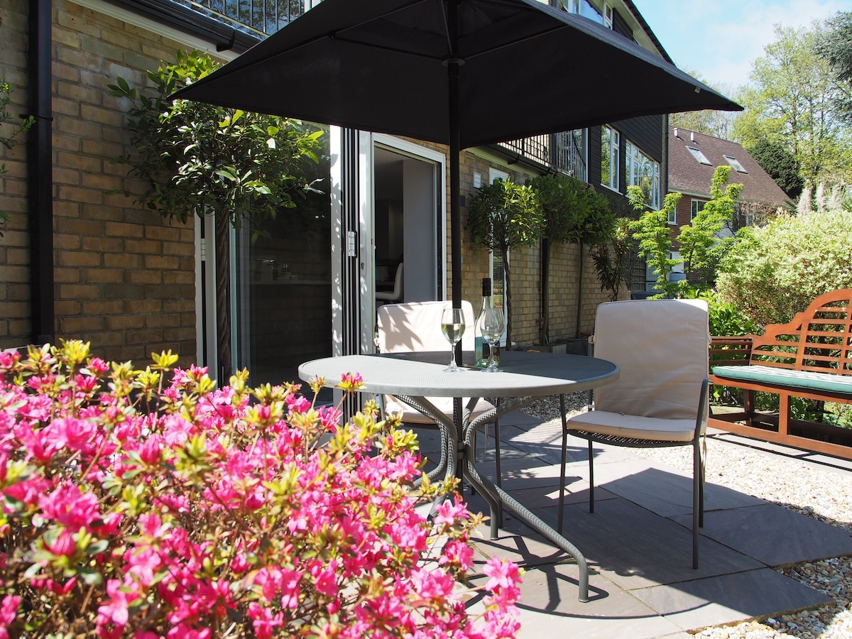 A garden seating area features a round table with two chairs under a dark umbrella, offering shade. Bright pink azaleas surround the space, adding color. In the background, doorways lead into the apartment, with greenery visible in the well-maintained garden.