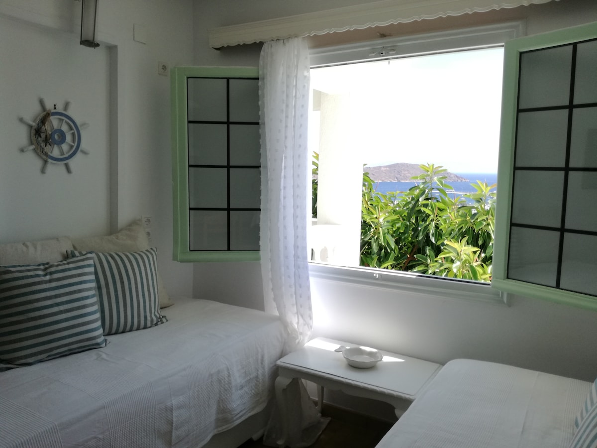 A well-lit room features two beds separated by a small table. Light curtains frame a window that opens to a view of greenery and the sea beyond. Decorative pillows add a touch of comfort to the bedding.