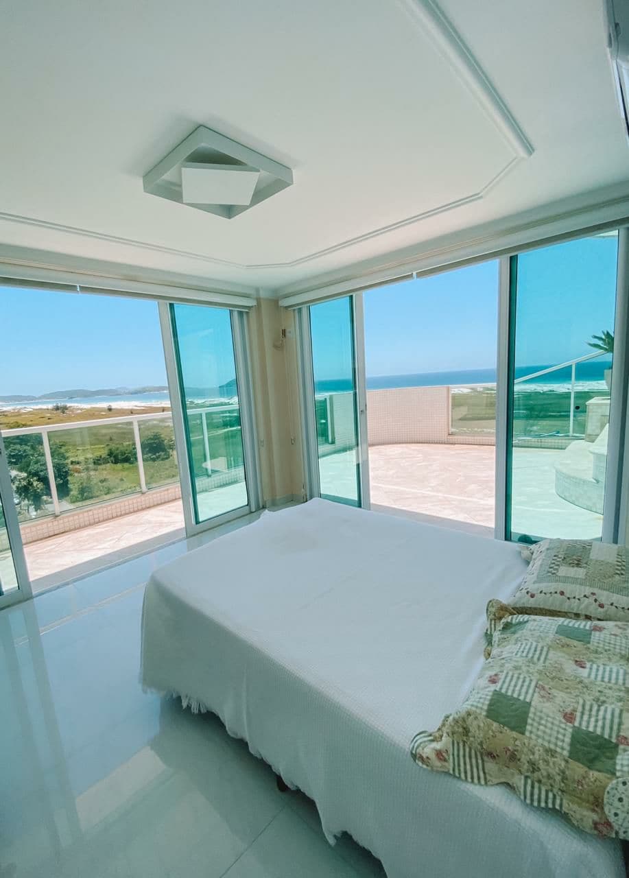 A light-filled bedroom features floor-to-ceiling windows offering panoramic views of the surrounding landscape and ocean. A spacious bed is positioned centrally within the room. Soft, neutral tones are complemented by a decorative throw at the bed's foot, enhancing the serene environment.