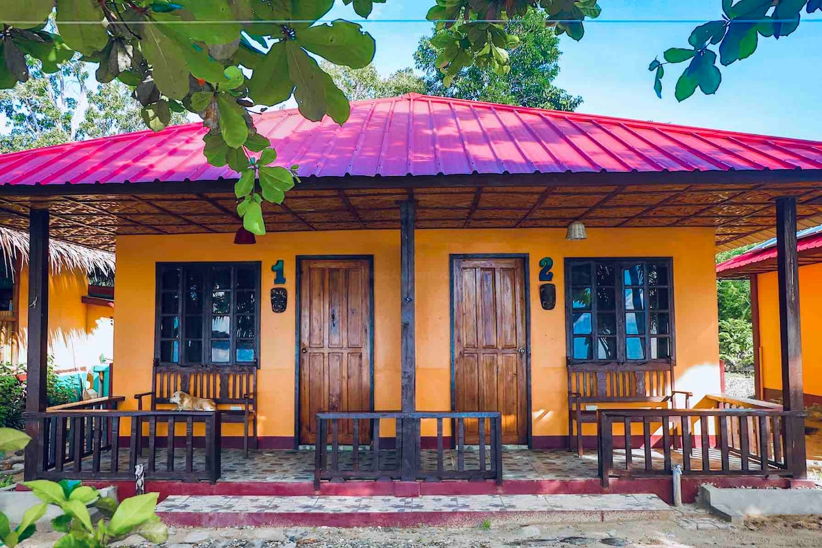 Two inviting cabins share a vibrant orange exterior, each featuring a wooden door and a small porch area with seating. A red metal roof offers shade, while green foliage frames the scene, creating a welcoming entrance for guests.