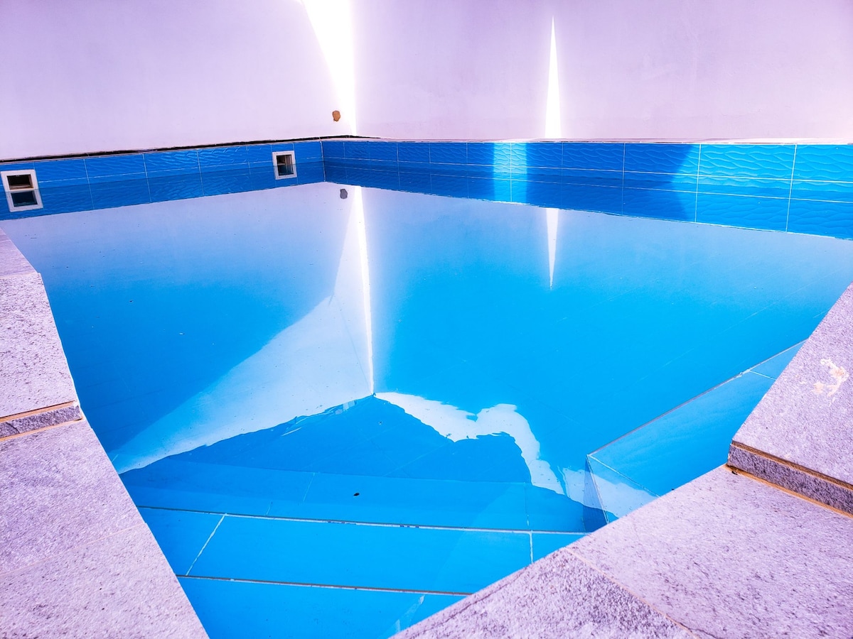 A clear swimming pool is set within a modern enclosure, featuring tranquil blue tiles. Sunlight reflects off the water's surface, creating soft light patterns across the pool. Surrounding edges are lined with light-hued stone, enhancing the overall serene atmosphere.