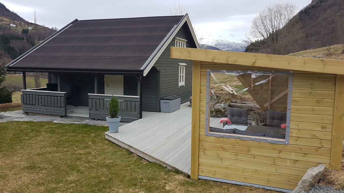 A modern cabin features a dark exterior with a spacious deck, complemented by a separate wooden lounge area with large windows. The landscape offers grassy areas and distant mountains, creating a serene backdrop. Comfortable seating is visible within the transparent-walled lounge.