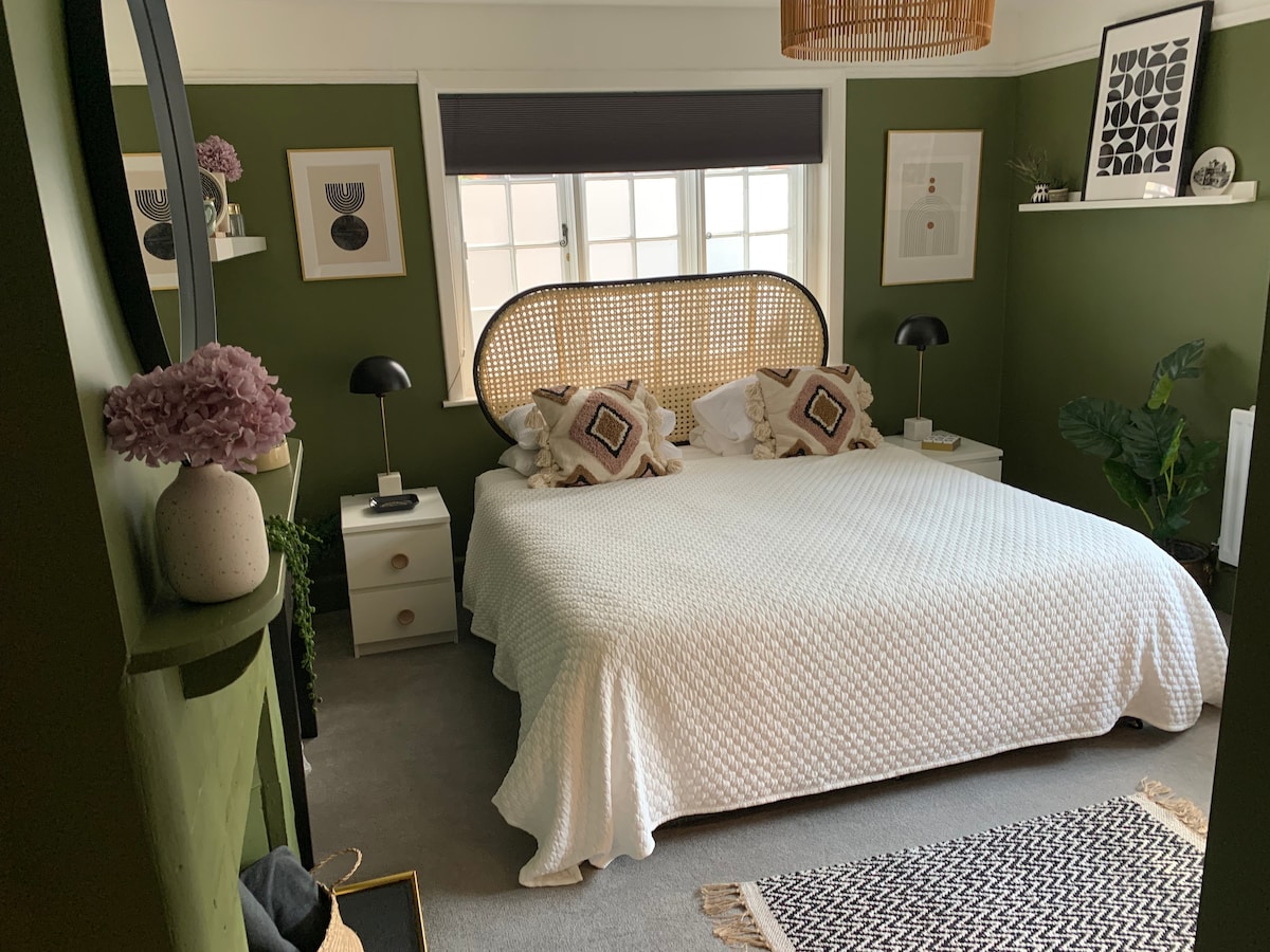 A spacious bedroom features dark green walls and a textured white bedspread draped over a large bed. Coordinating lamps sit on bedside tables adorned with decorative wall art. A rug adds contrast to the floor, while natural light filters through a window with shades.