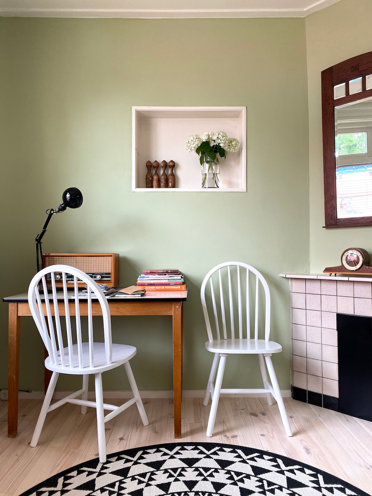 A small living area is displayed with a light green wall and a decorative niche featuring a vase of flowers. A wooden table is accompanied by two white chairs, with a vintage record player and books on the table. A patterned rug adds texture to the space.