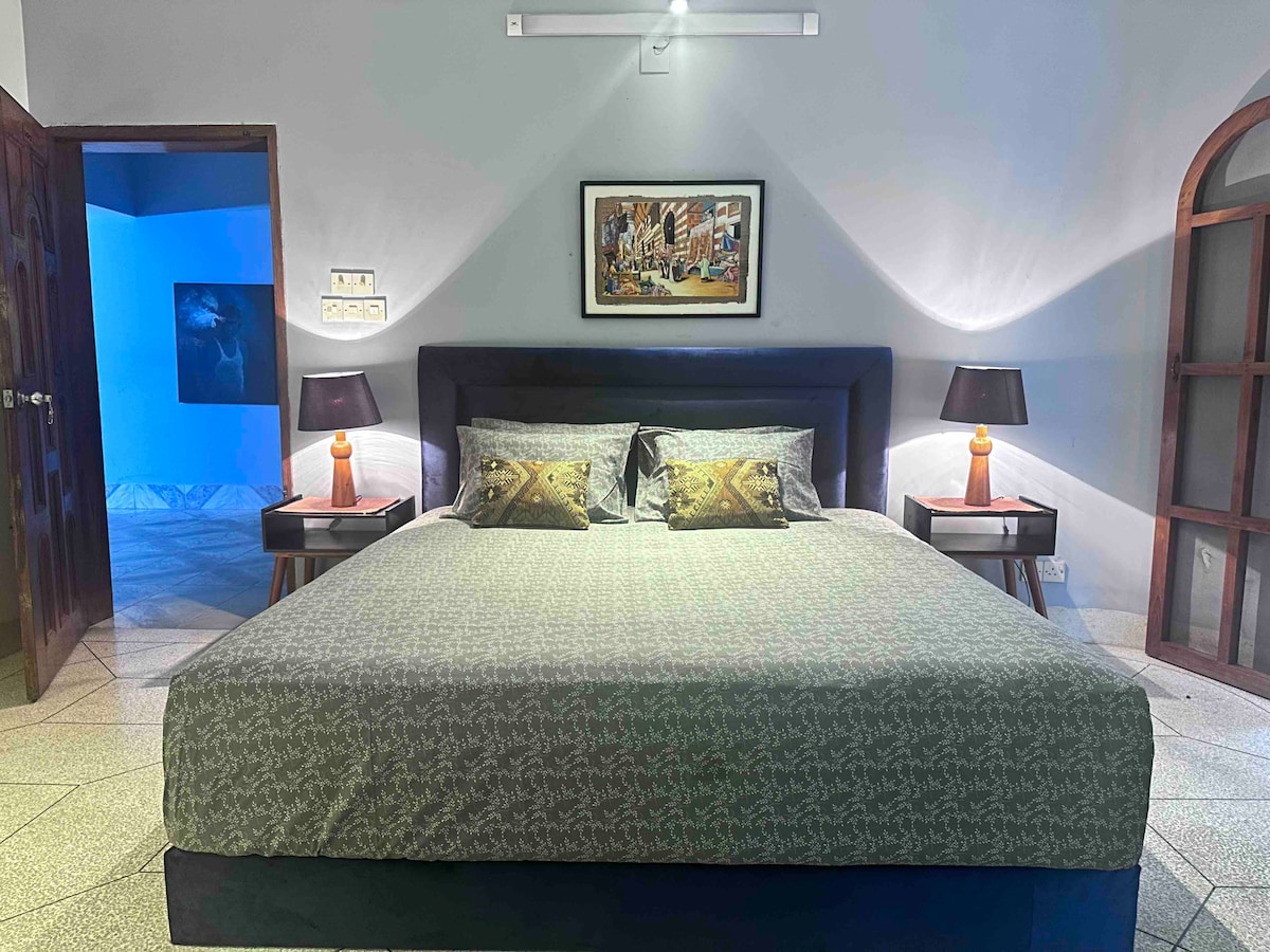 A spacious bedroom is featured, furnished with a large bed adorned with a patterned green cover. Flanking the bed are two bedside tables, each with a lamp casting soft light. A framed artwork hangs above the bed, complementing the room's decor.