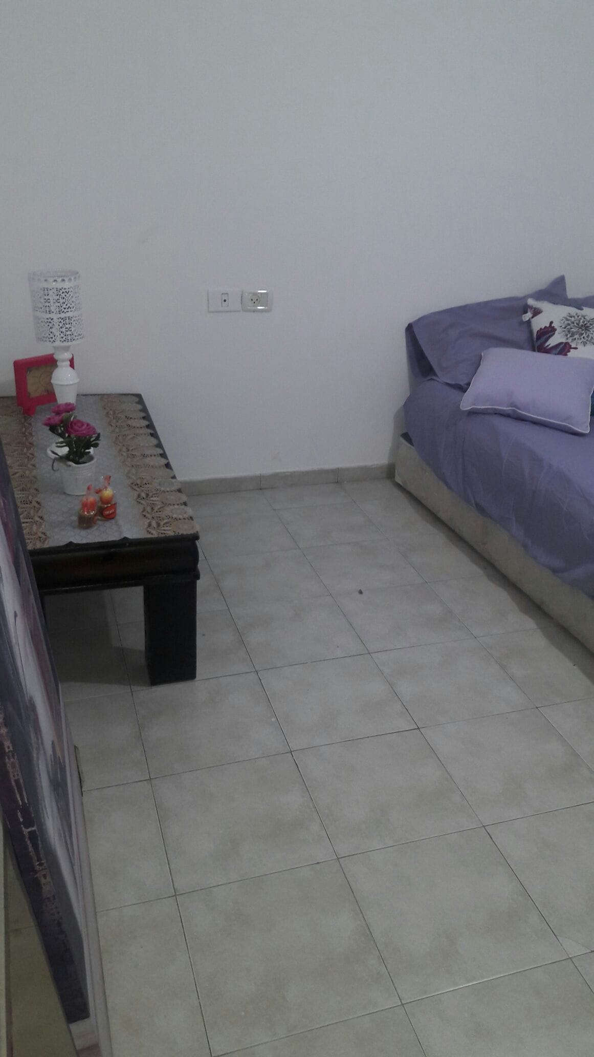 A cozy sleeping area is depicted, featuring a spacious bed covered with light purple linens and decorative pillows. A simple bedside table holds candles and a small decorative piece. Light-colored tiles cover the floor, contributing to the room’s bright atmosphere.