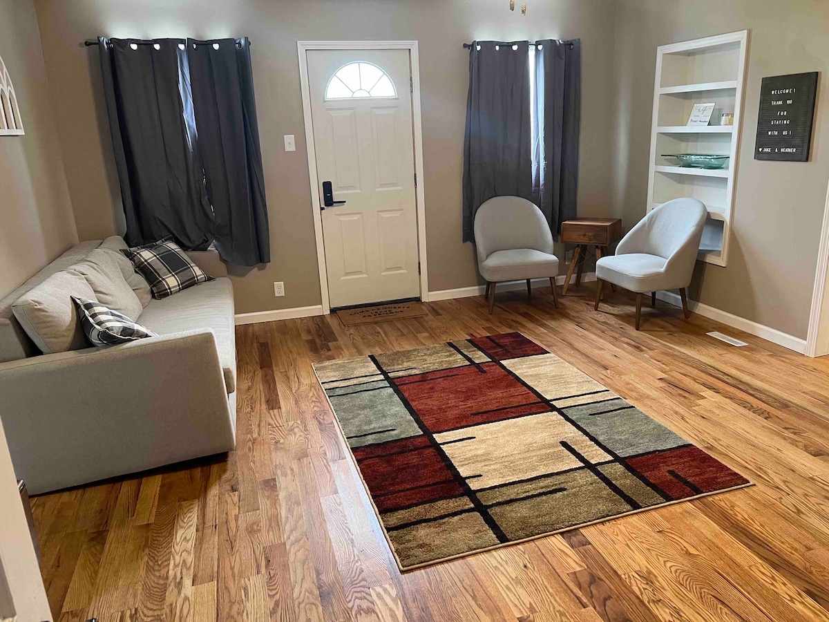 The living area features hardwood flooring and is furnished with a comfortable couch and two accent chairs. A decorative area rug with geometric patterns adds color, while natural light is diffused through the sheer curtains at the window. The front door is visible, leading outside.