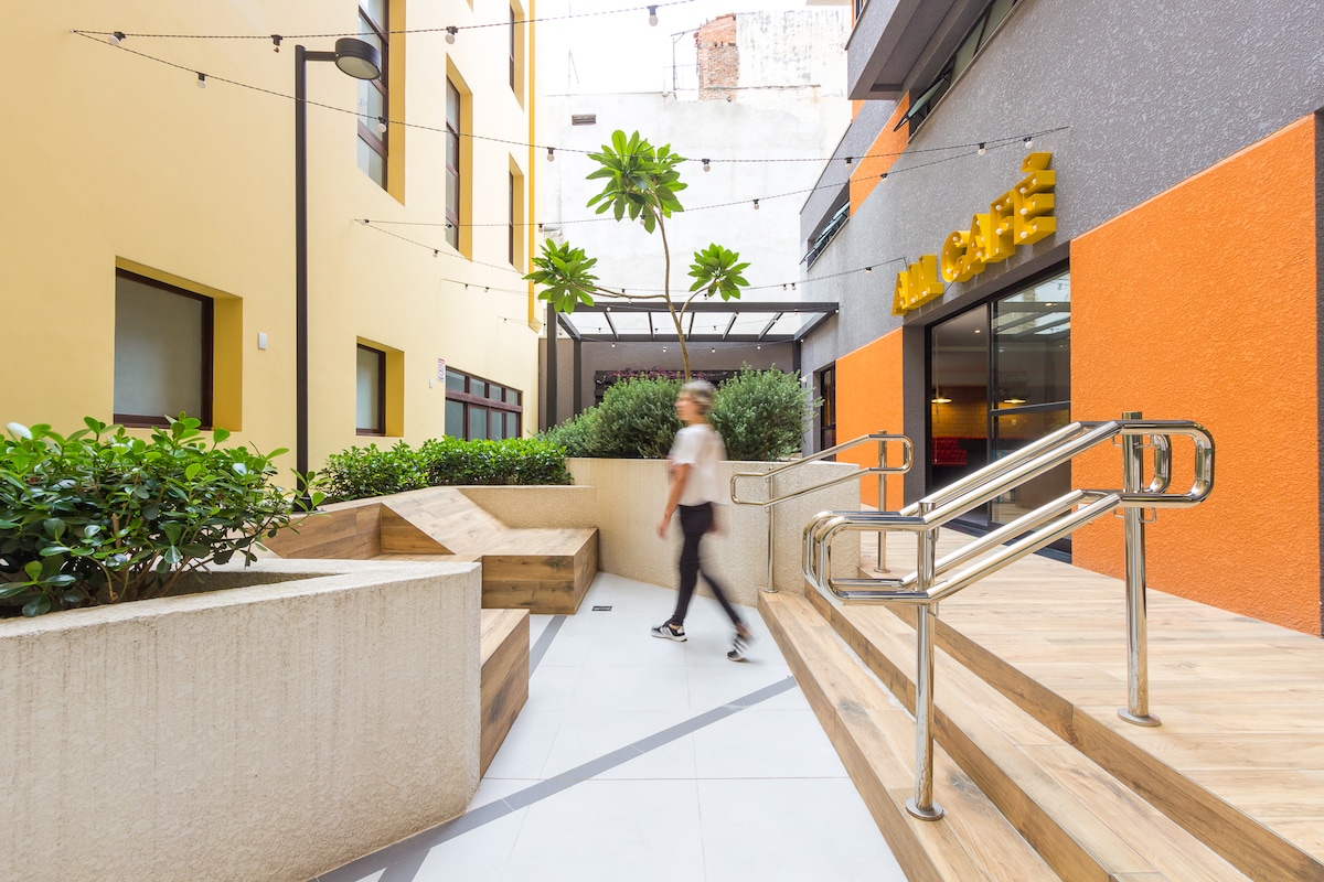 An inviting outdoor area showcases a modern café with bright orange accents. Wooden seating and lush greenery create a relaxing ambiance, while accessible pathways and handrails ensure convenience for all guests. The space is bathed in natural light, providing a welcoming environment.