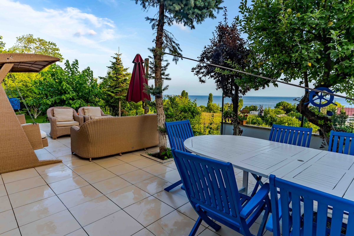 An outdoor patio area features a large white dining table surrounded by blue chairs, accommodating multiple guests. A shaded seating arrangement is provided, accompanied by lush greenery and a view of the nearby sea. The tiled floor enhances the spacious feel of this inviting setting.