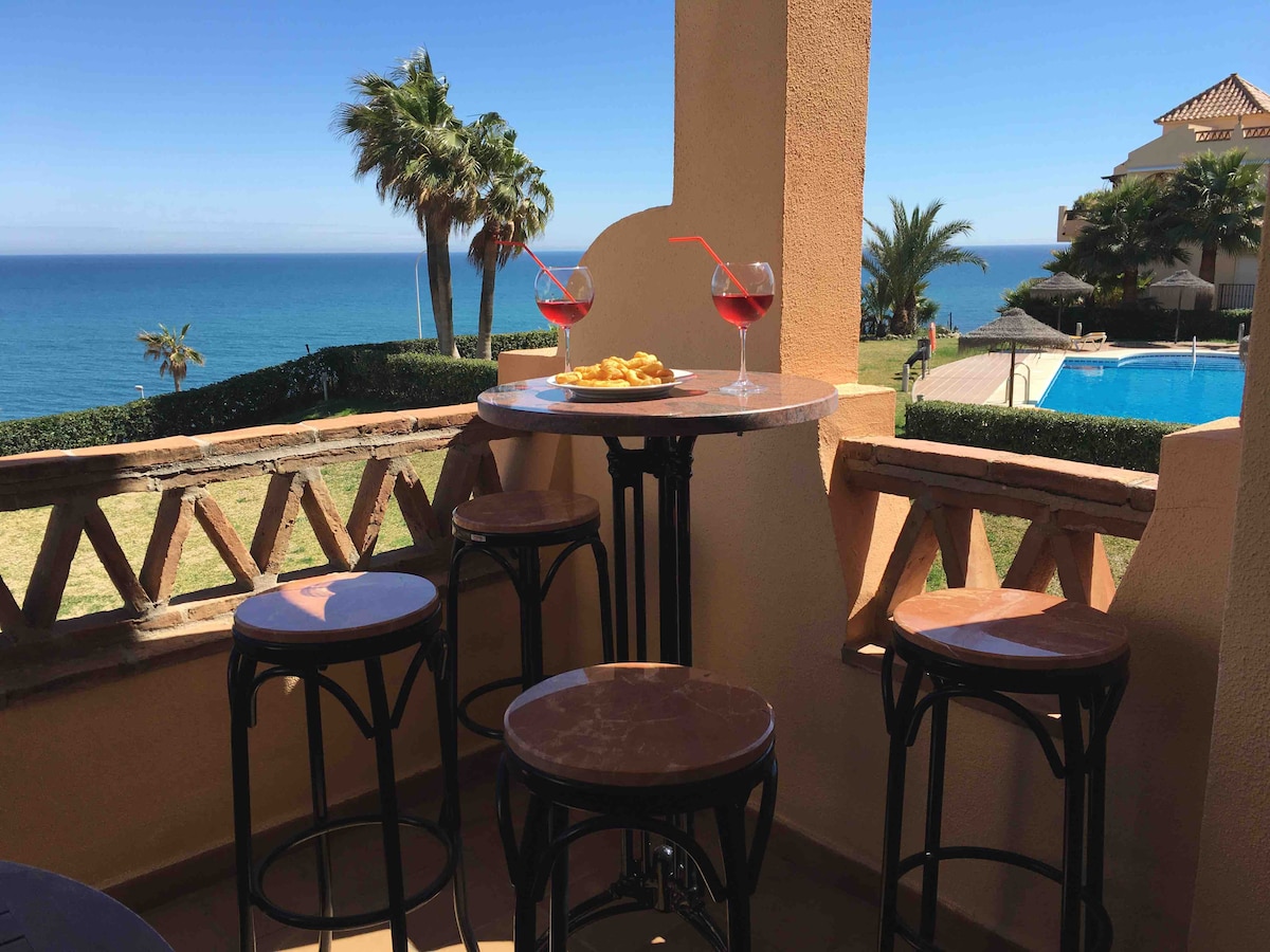 A cozy terrace features a marble-top table surrounded by four tall stools. Two drinks are placed on the table, accompanied by a plate of snacks. The view includes palm trees and a blue sea, creating a serene backdrop.
