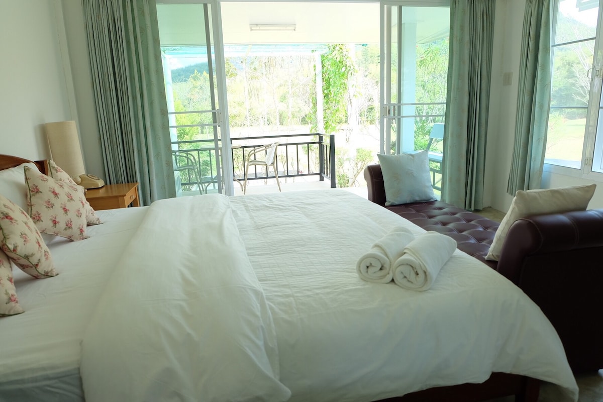 A bright bedroom is designed with a spacious bed adorned with floral pillows. Natural light filters in through large glass doors, leading to a balcony with views of lush greenery. A cozy seating area includes a plush chair, complemented by rolled towels arranged neatly.