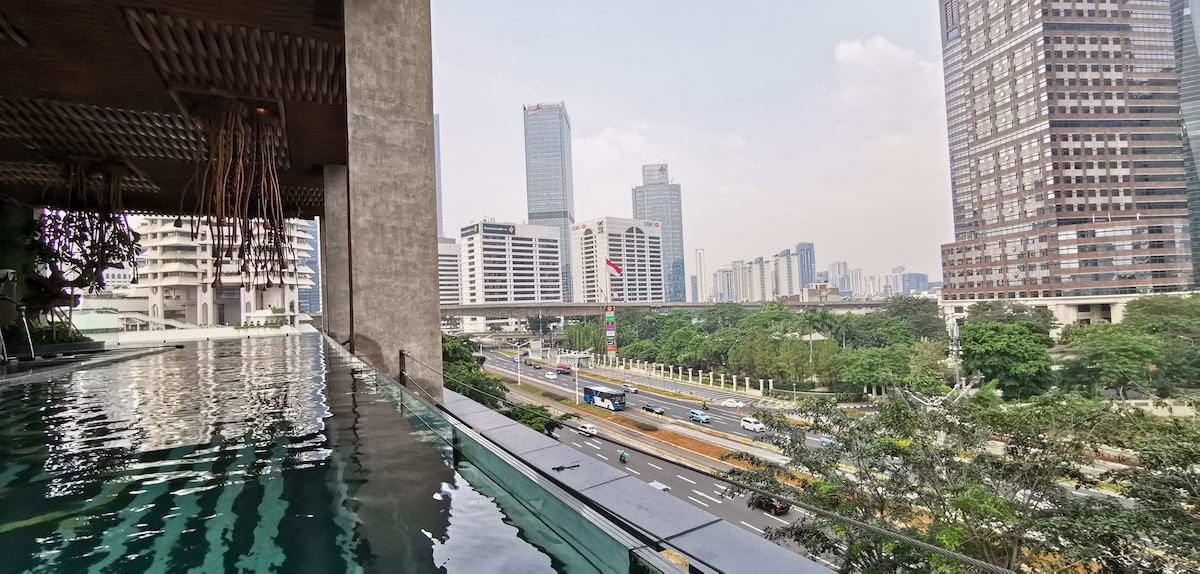 A view from the pool area showcases the bustling Sudirman road below, with tall buildings lining the skyline. A reflection of the pool's edge can be seen, enhancing the connection between the interior and the vibrant cityscape.