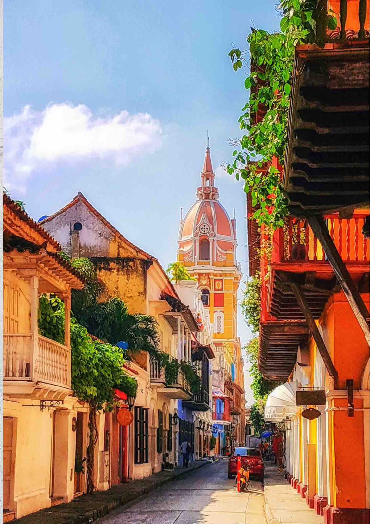 A narrow street is lined with traditional colonial buildings, showcasing vibrant colors and lush greenery. At the end of the street, a tall clock tower stands prominently, surrounded by a clear blue sky with scattered clouds.
