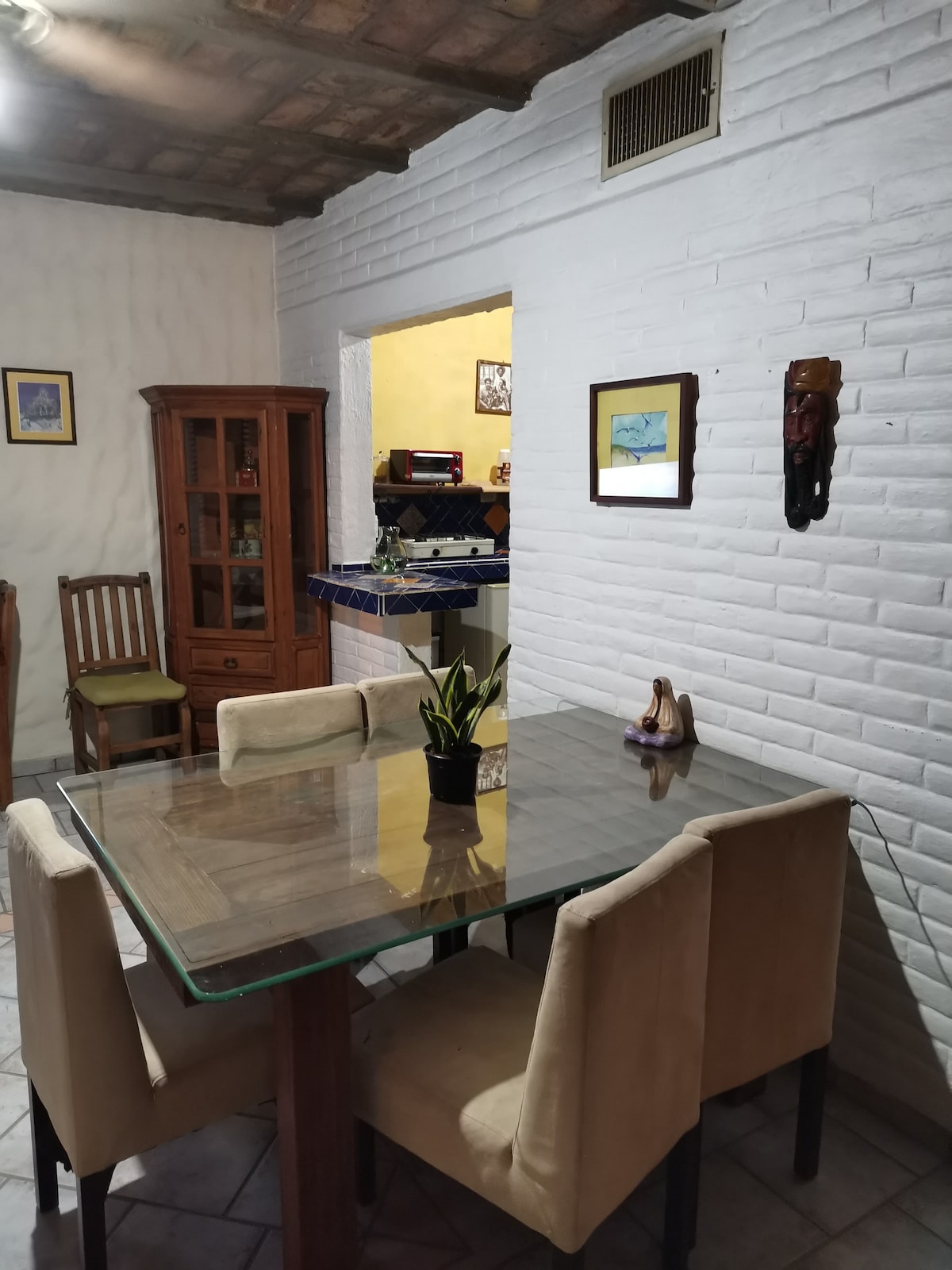 A dining area features a glass-top table surrounded by four cushioned chairs. A wooden cabinet is positioned against a white-brick wall, while decorative artwork frames the space. An open doorway leads to the kitchen, visible in the background.