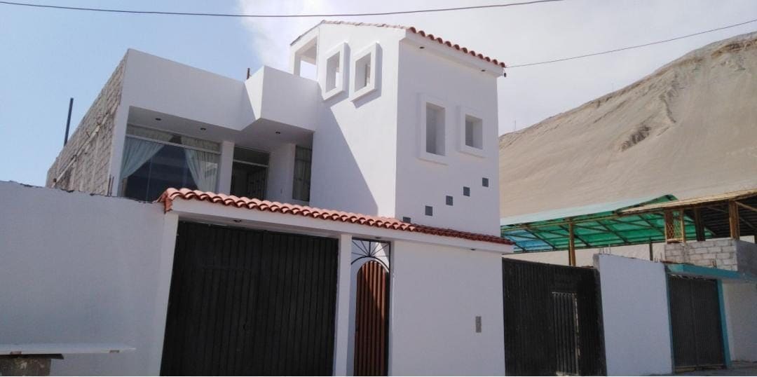 A modern two-story house displays a clean white facade with minimalist design elements. Large windows and a unique rooftop feature are visible, while a traditional red-tiled roof provides a contrasting touch. A wrought iron gate and the surrounding landscape are also present.