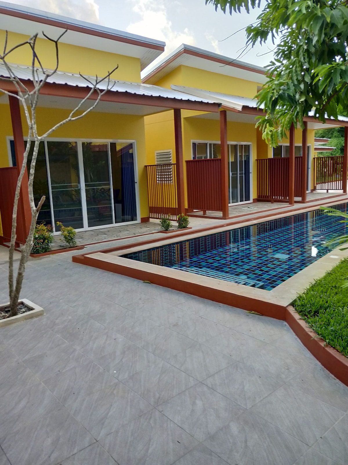 A serene outdoor area is highlighted by a rectangular swimming pool featuring a vibrant tile design. The surrounding space is bordered by neatly maintained gardens and wooden fences. Yellow walls of the nearby structures create a bright and inviting environment.