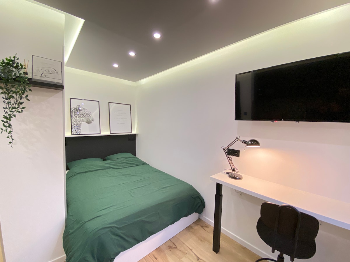 A spacious sleeping area features a double bed covered with a dark green comforter. Above the bed, two framed artworks are displayed. A wall-mounted television is visible, alongside a desk with a modern lamp and a black chair. Soft lighting enhances the room's ambiance.