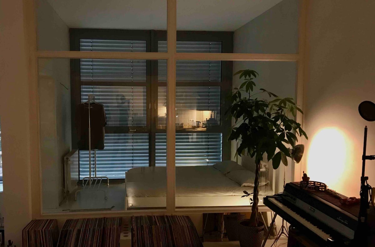 The bedroom is designed as a glass-walled space, showcasing a spacious bed and a potted plant nearby. A garment rack can be seen, and soft lighting from nearby windows creates a serene environment, while minimalistic furniture accents the modern aesthetic.