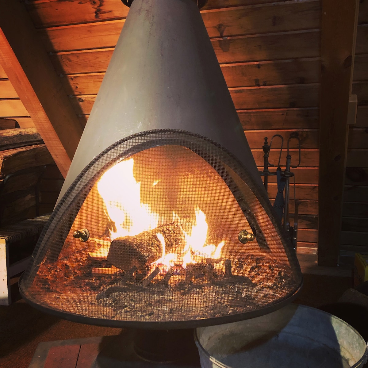A modern fireplace features a cone-shaped metal structure with a mesh screen, showcasing a warm fire burning within. Logs are arranged on the grate, surrounded by a bed of ash. Nearby, accessories for maintaining the fire are visible.
