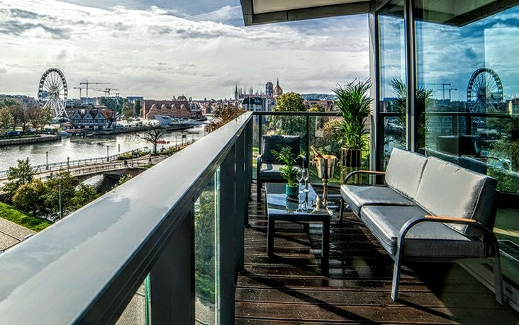 An outdoor terrace features comfortable seating and a glass table, overlooking a scenic view of the river and the historic city. Nearby, a Ferris wheel is visible, contributing to the vibrant surroundings filled with greenery.