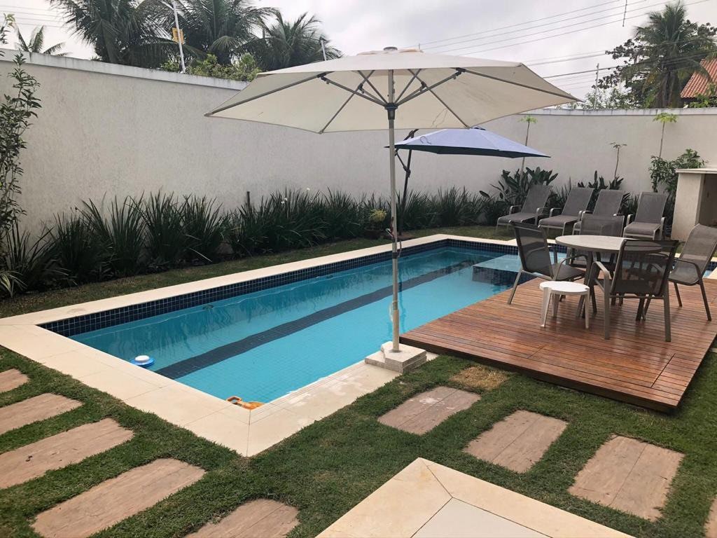 A well-maintained outdoor space features a serene swimming pool surrounded by lush greenery. Lounge chairs and an umbrella provide shaded seating on a wooden deck, while a dining table with multiple chairs offers a spot for outdoor meals.