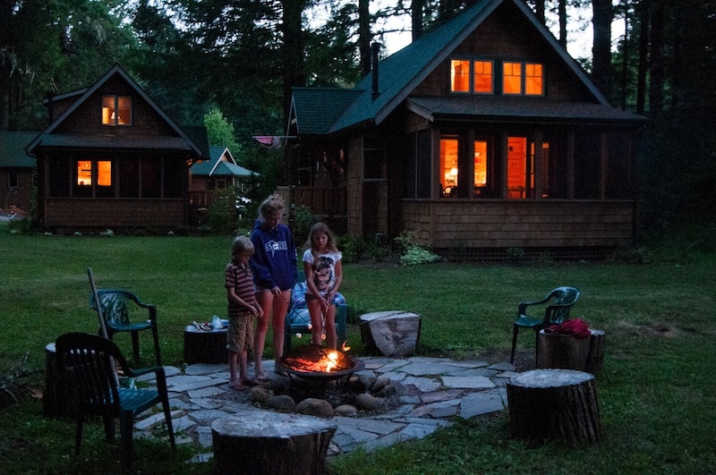 A serene fire pit is surrounded by log seating, with children standing nearby. Two cabins with illuminated windows are visible in the background, nestled among trees. The grassy area provides space for outdoor activities, enhancing the natural ambiance of the setting.