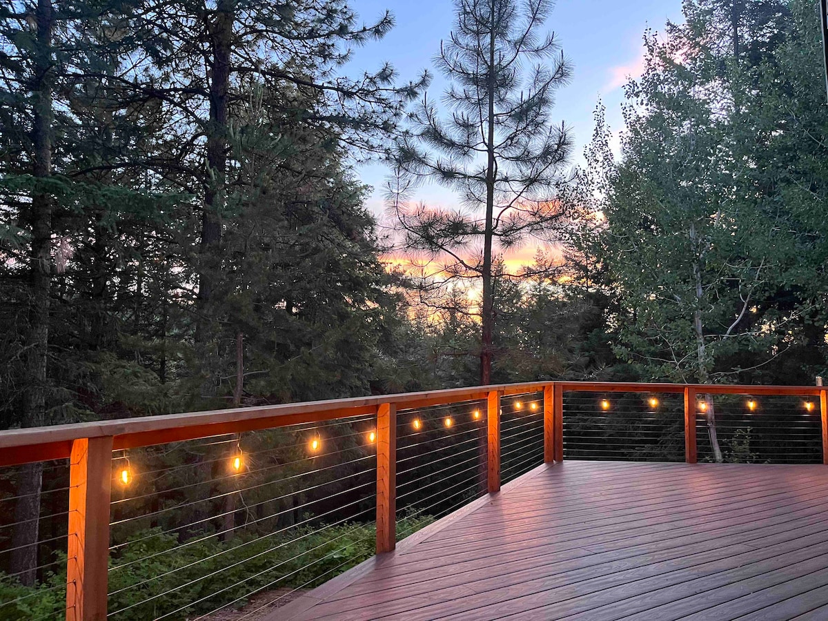 A large wooden deck is surrounded by tall trees, offering a peaceful view of the colorful sunset in the distance. Soft lighting is provided by string lights along the railing, creating a warm ambiance. Natural greenery surrounds the deck, enhancing the secluded feel.