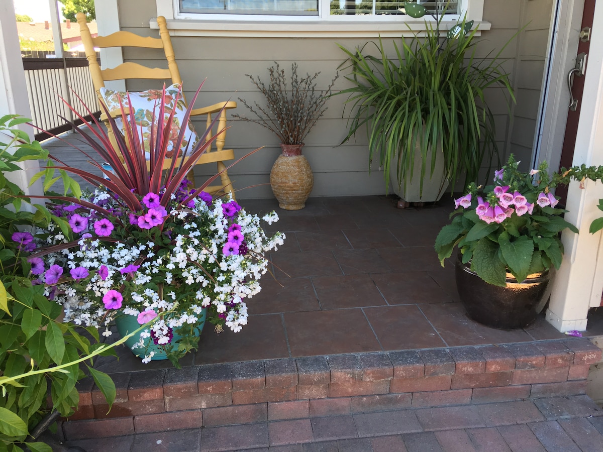 A welcoming front porch is adorned with vibrant potted flowers in shades of purple and white. A rustic chair sits beside a tall potted plant, complementing the overall charm. Textured planters hold additional greenery, creating an inviting atmosphere for relaxation.