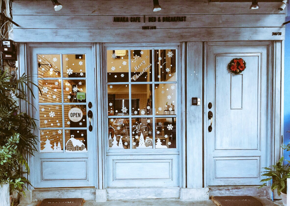 The exterior of Amaka Cafe & Bed & Breakfast features a charming blue façade adorned with decorative snowflakes and a festive wreath. The open sign invites guests to enter, while the windows showcase a cozy atmosphere within the café, hinting at the welcoming environment.