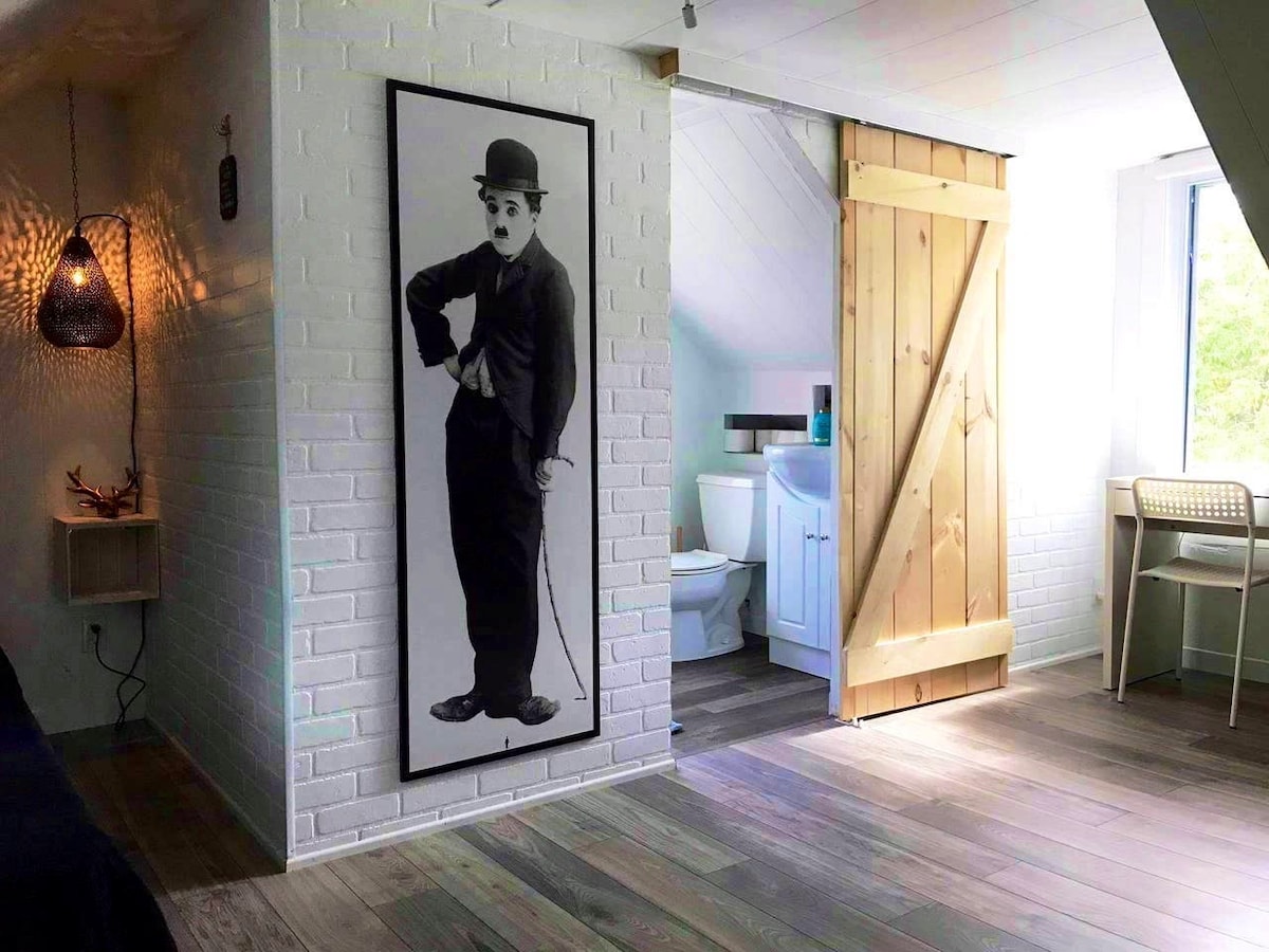 A stylish loft space features a striking black-and-white portrait on a white brick wall. A wooden sliding door leads to a bathroom area. Natural light floods the room through a large window, illuminating the modern decor and wooden flooring.