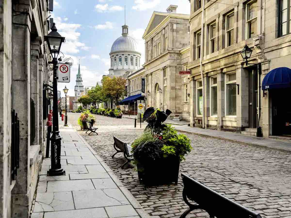 A charming cobblestone street is bordered by historic buildings, featuring flower pots filled with vibrant plants. Benches are positioned along the walkway, with lamp posts guiding the path. The domed structure in the background marks a notable landmark, enhancing the ambiance of the area.