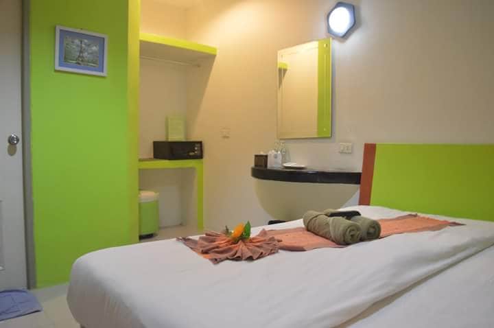 A brightly colored room features a spacious bed dressed in white linens. A decorative towel arrangement sits at the foot of the bed. A compact seating area and a mirror are visible, alongside amenities such as a kettle and cups. Soft lighting enhances the vibrant green walls.