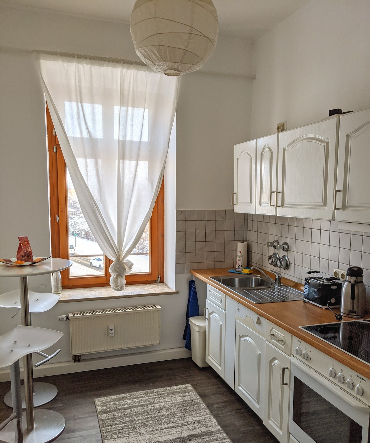 A well-equipped kitchen features white cabinetry and wooden countertops. A window draped with light fabric allows natural light to enter. A round dining table with modern stools is visible in the corner, along with essential appliances including a coffee maker and stove.