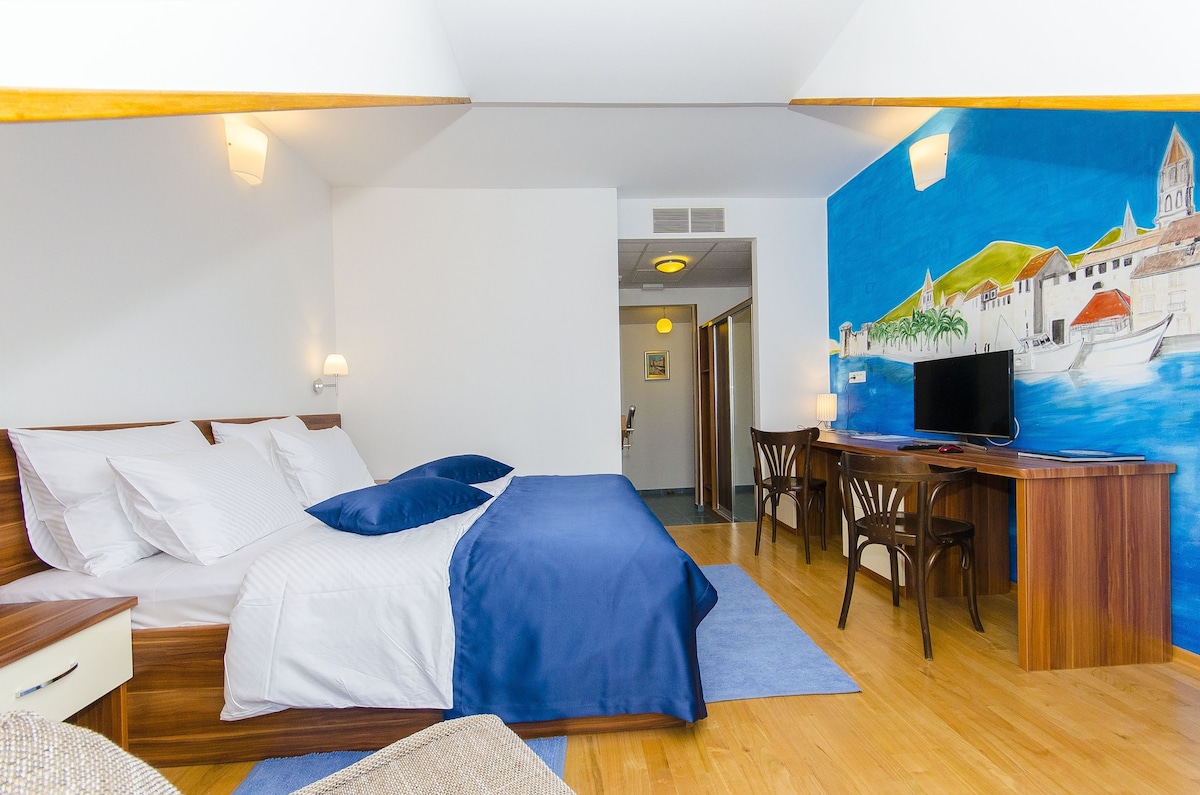 The spacious room features a large bed dressed in white linens, complemented by a blue throw. A mural depicting a coastal scene serves as a vibrant backdrop. A wooden desk and chair are positioned near a television, with warm light fixtures illuminating the area.