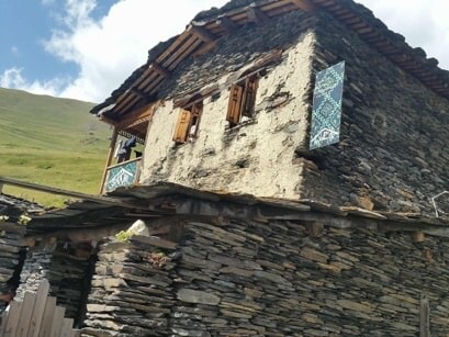 An exterior view of a rustic stone house is presented, featuring a weathered facade and wooden window frames. Intricate designs adorn the window shutters, adding character. The surrounding landscape includes green hills and a partly cloudy sky, creating a serene backdrop.