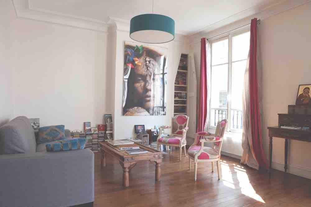A bright living room features a spacious sofa and two elegant chairs against a backdrop of large windows. A wooden coffee table occupies the center, surrounded by bookshelves filled with books and decorative items. Natural light filters in through the windows, enhancing the room's inviting feel.