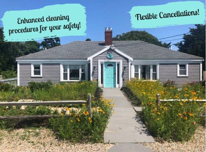 The exterior of a single-level beach house is presented, featuring grey shingle siding and a turquoise front door. A pathway leads from the entrance, bordered by colorful wildflowers. A fence provides framing against the green landscape and a clear blue sky.