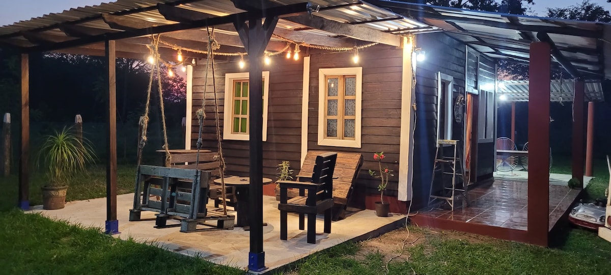 A quaint outdoor seating area is featured under a metal roof, illuminated by soft lights. Wooden furniture, including a swing and chairs, surrounds a rustic table. Lush greenery and potted plants complement the space, providing a natural and inviting setting for relaxation.