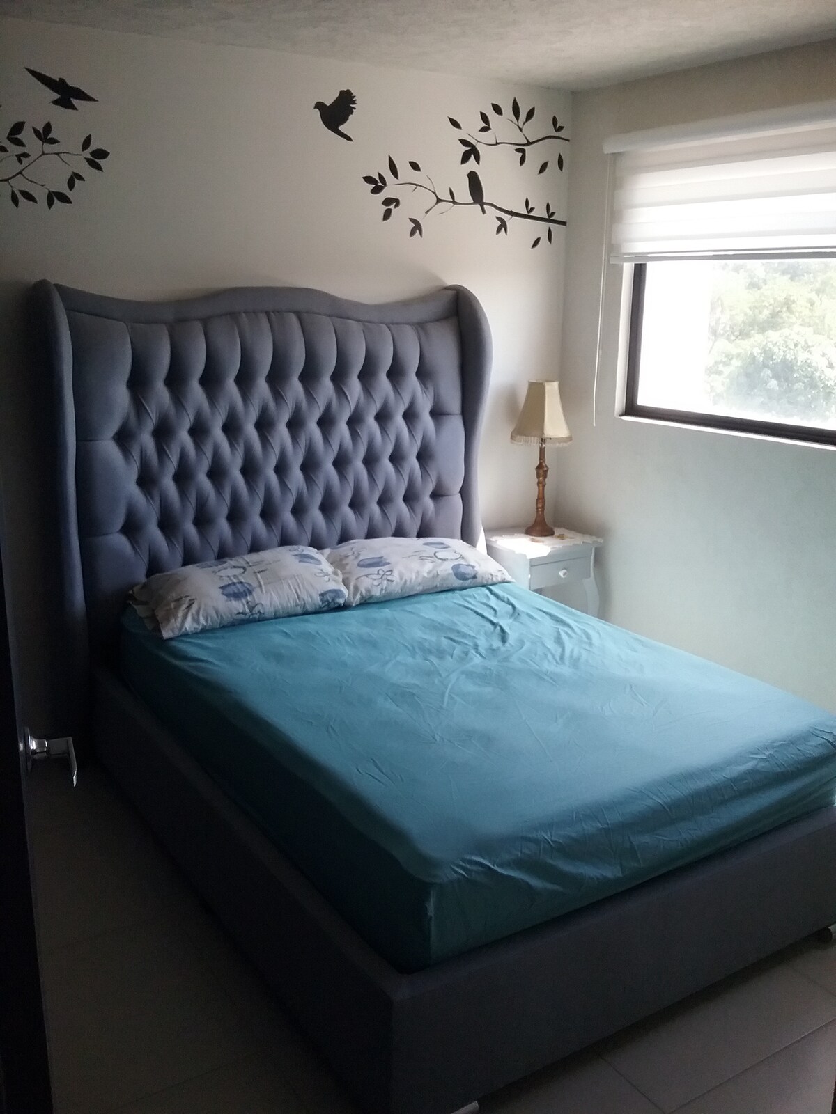 A private bedroom features a spacious bed with a light blue fitted sheet and patterned pillows. Decorative wall decals of birds and branches enhance the ambiance. Natural light enters through a large window covered with a sleek blind, contributing to a calm atmosphere.