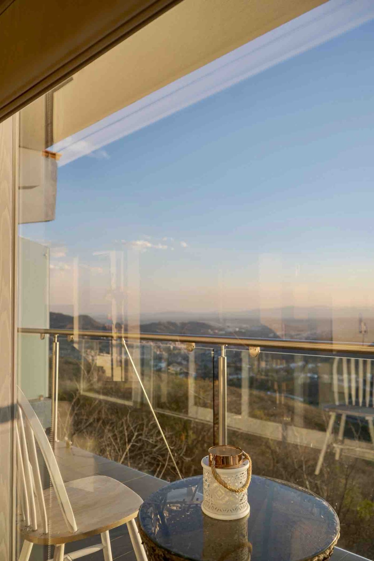 A balcony features a small round table and a white chair, offering a space to enjoy the view. Glass doors reflect the landscape beyond, with soft evening light casting a gentle glow on the surroundings.
