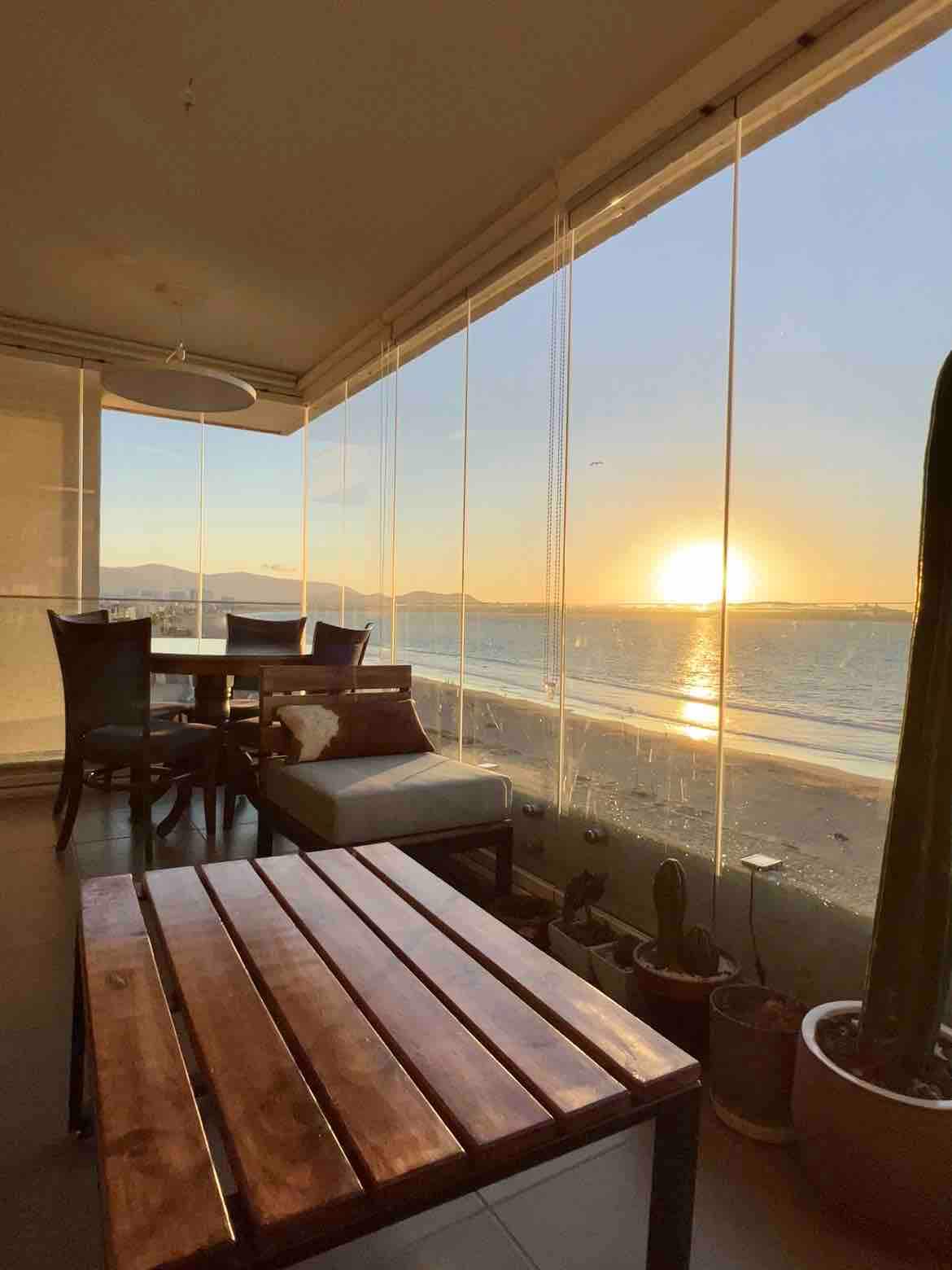 A well-lit terrace boasts a wooden table and chairs, offering an unobstructed view of the ocean and sunset. Floor-to-ceiling glass windows allow natural light to fill the space, enhancing the tranquil atmosphere. Potted plants are placed along the edges, adding greenery.