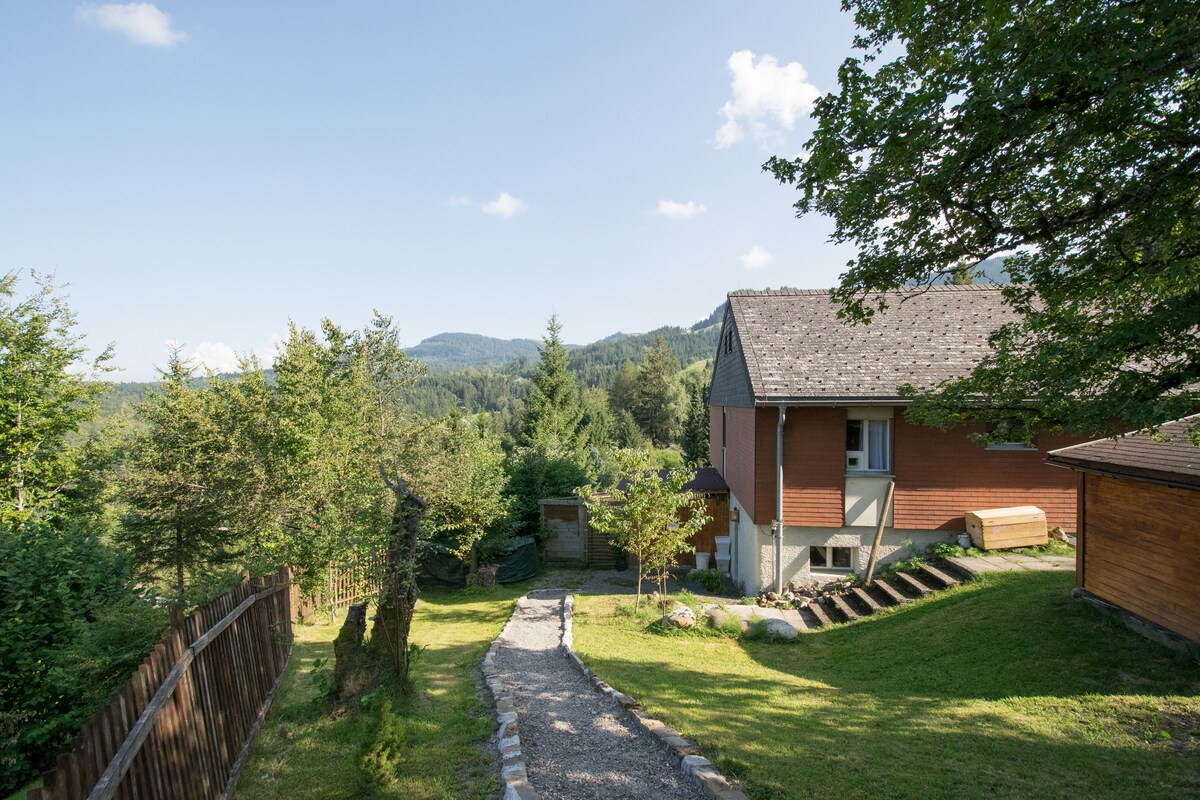 A scenic view of the property is presented, featuring a path leading from a fenced garden toward the house. Surrounding greenery includes trees and shrubs, with gentle hills visible in the background under a clear blue sky. The structure shows a mix of wood and stone materials.