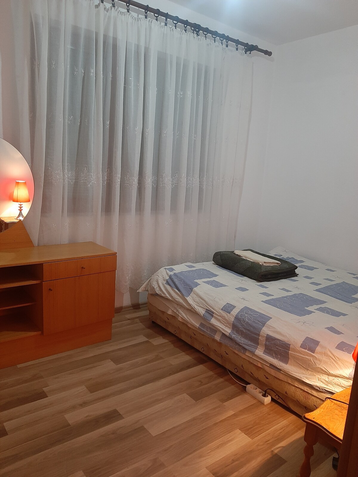 A simple bedroom features a spacious bed covered with a patterned blanket and a folded towel. A wooden nightstand with a lamp is positioned beside the bed. Natural light filters through sheer curtains, illuminating the room's light-colored walls and wooden flooring.