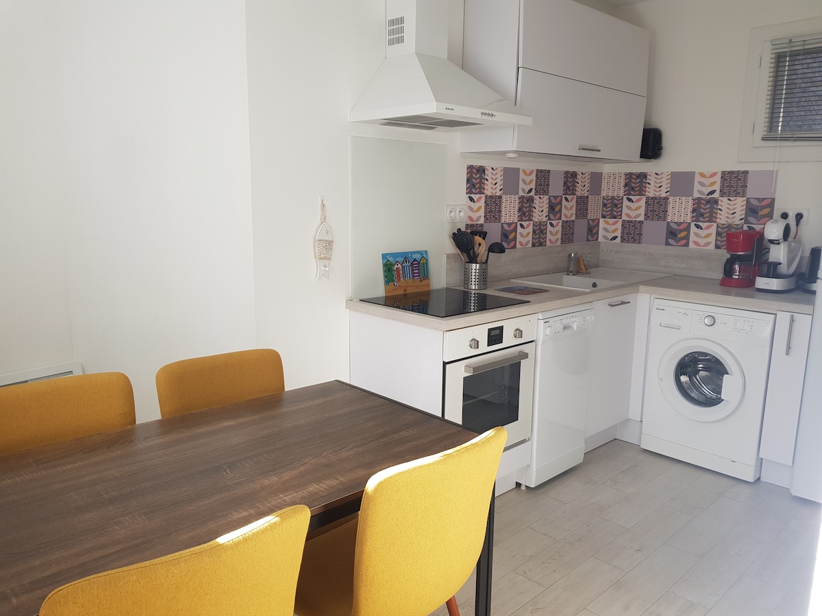 A well-equipped kitchen with white cabinetry and colorful patterned backsplash is featured. A wooden dining table is surrounded by four yellow chairs. Appliances include a gas stove, oven, dishwasher, microwave, and washing machine, alongside various cooking utensils and a coffee maker.