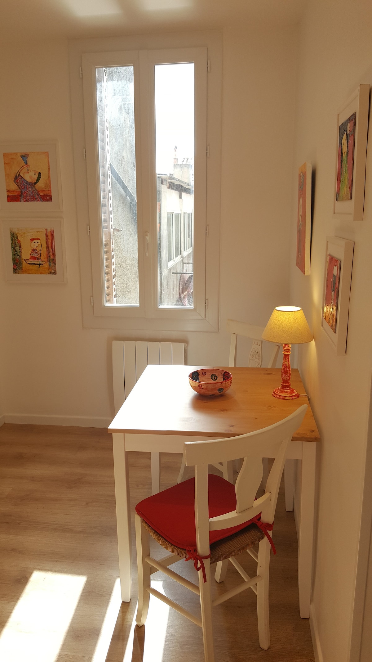 A small dining area features a light-colored wooden table accompanied by two white chairs, one with red cushions. A bowl sits at the center of the table. The space is illuminated by a lamp, and colorful artwork adorns the walls, adding visual interest.